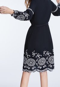 Robe noire à manches longues bouffantes, ornée de broderies florales blanches aux poignets et à l'ourlet, taille cintrée, texture lisse, ourlet arrondi.