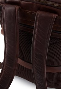 Brun læderrygsæk med polstret netpåsyet rygstøtte, dobbelte stropper og en lynlåslomme. Har tekstureret læder og kontrasterende syninger.