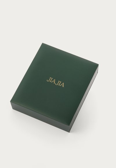 Zaļš, spīdīgs iepakojuma kastīte ar kvadrātu formu un zelta burtiem "JIA JIA" augšpusē. Gluda tekstūra bez papildu zīmējumiem vai akcentiem.