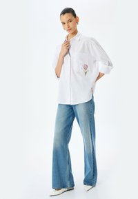 Camicia bianca con bottoni e tasca, decorata con ricami di tulipani rosa; abbinata a jeans a gamba larga blu e scarpe bianche a punta.