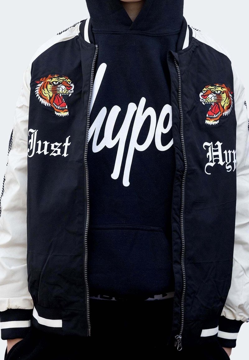 Veste bomber noire avec manches blanches, ornée de motifs brodés de tigres et de textes. En dessous, un sweat à capuche noir avec de grandes lettres blanches.