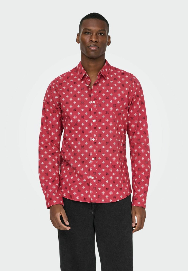 Man in een rood overhemd met knopen en lange mouwen, versierd met witte en zwarte sterpatronen, in combinatie met een zwarte broek, staande tegen een eenvoudige lichte achtergrond.