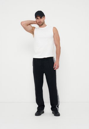 Férfi fekete sapkát, ujjatlan fehér inget, fekete fehér csíkkal díszített melegítőnadrágot és fekete cipőt visel, egy kézzel a nyaka mögött áll.