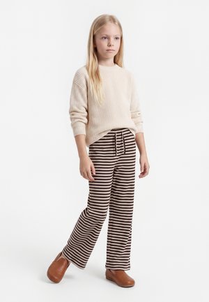Jeune fille debout portant un pull côtelé crème, un pantalon large rayé marron et beige, et des chaussures marron sans lacets sur fond blanc.