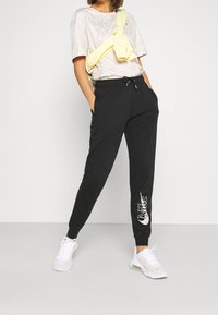 Svarta sweatpants med elastiskt midjeband och sidofickor, med en vit Nike-logotyp tryckt på vänster ben. Kombineras med en ljus grafisk t-shirt.