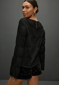 Schwarzes, strukturiertes Langarmoberteil mit Rundhalsausschnitt und Schlüssellochdetail am Rücken, kombiniert mit schwarzen Samtshorts.