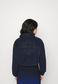 Ναυτικό κοντό σούπερ-τούπ με ψηλό γιακά, υφασμάτινη υφή, ανάγλυφο λογότυπο "adidas ATHLETIC LEAGUE" στην πλάτη και ριμπ μανίκια.