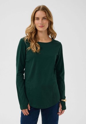 Jeune femme aux longs cheveux blonds ondulés, portant une chemise à manches longues vert foncé et un jean bleu, debout devant un fond uni.