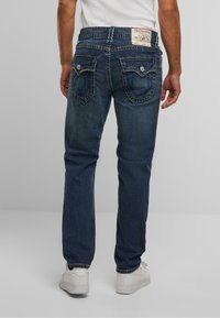 Jeans de mezclilla azul oscuro con corte recto, bolsillos traseros visibles con diseños cosidos y una etiqueta de cuero de marca en la cintura.