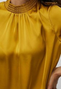Top giallo senape con scollo smock, dettagli plissè e una texture leggera e liscia. Design elegante, semi-adattato.