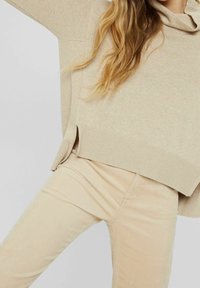 Person som bär en ljusbeige stickad tröja med vida ärmar och matchande kordbyxor mot en neutral bakgrund.