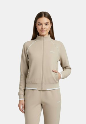 Donna in piedi con una mano nella tasca della sua giacca beige con cerniera, indossa pantaloni sportivi beige abbinati su uno sfondo semplice.