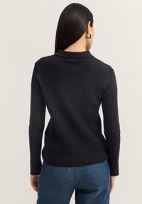 Schwarzer Rippstrickpullover mit Kragen und langen Ärmeln. Der Stoff wirkt weich, strukturiert und körperbetont, kombiniert mit blauen Jeans.