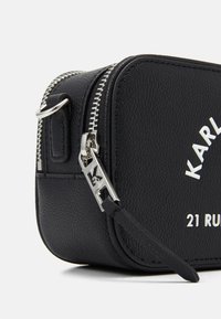 Czarna skórzana prostokątna torba z srebrnymi elementami, zamknięciem na zamek błyskawiczny i logo. Posiada teksturowaną powierzchnię oraz boczny pasek.