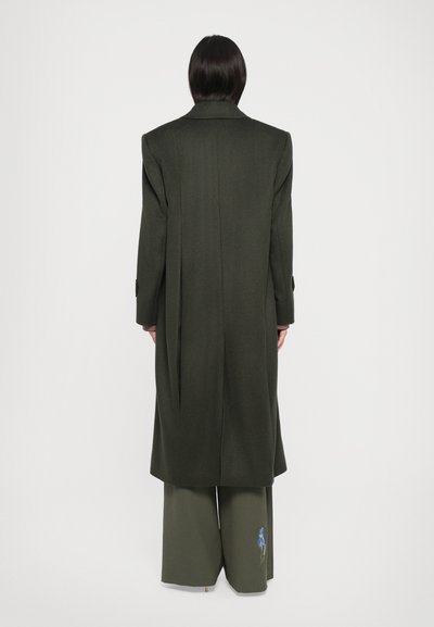 Long manteau en laine vert olive avec une silhouette structurée, de larges épaules et des fentes sur les côtés, porté sur un pantalon large assorti orné de broderies florales.