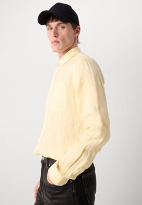 Light yellow button-up shirt with a single chest pocket, long sleeves, and textured fabric, paired with dark jeans. Black cap worn.