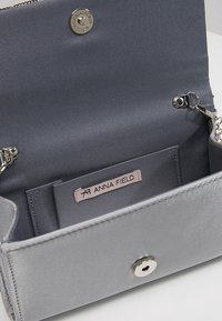 Šedý saténový interiér kabelky s kovovým uzávěrem na stisk, stříbrným řetízkem a štítkem s nápisem "ANNA FIELD." Uvnitř se nacházejí sloty na karty.