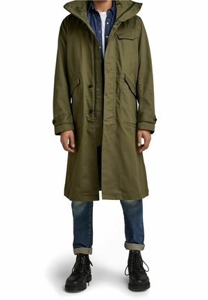 Homme portant un trench-coat à capuche vert olive, une chemise en jean bleue, un jean retroussé et des bottes noires à lacets, debout sur un fond blanc.
