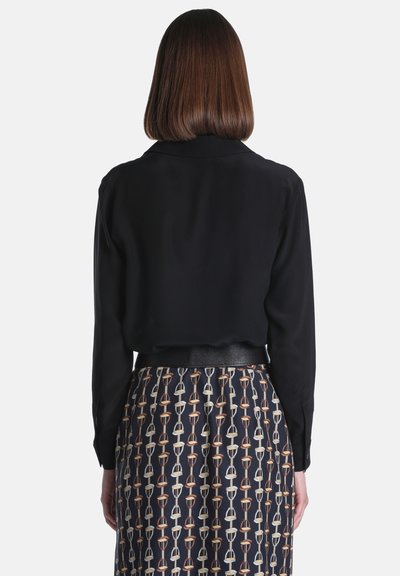 Femme vue de dos portant un haut noir à manches longues et une jupe avec un motif étrier marron et beige sur fond sombre.