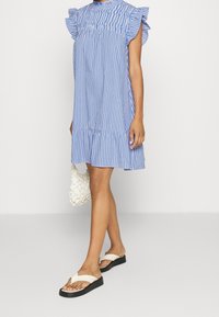 Robe rayée bleue et blanche avec des manches volantées, un col froncé et un ourlet à volants. Portée avec des sandales blanches et un sac beige texturé.