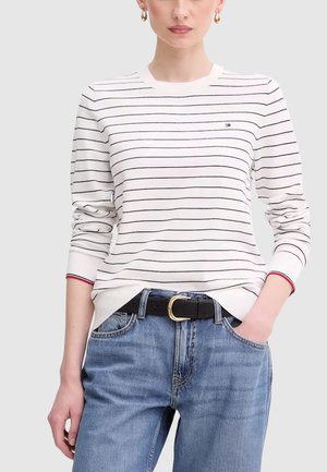 Donna che indossa una camicia bianca a maniche lunghe con strisce orizzontali nere e jeans blu, con la mano in tasca, su uno sfondo chiaro e uniforme.