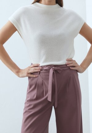 Vrouw draagt een witte gebreide top met korte mouwen en hooggesloten mauve broek met strik, staand met handen in de zij.