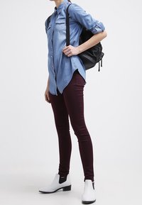 Ljus denimskjorta med knappar, maroon tight jeans och vita ankelskor med svarta accenter, kombinerat med en svart ryggsäck.