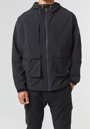 Mann trägt eine schwarze Kapuzenjacke mit Reißverschluss und großen Frontklappentaschen, kombiniert mit schwarzen Cargohosen.