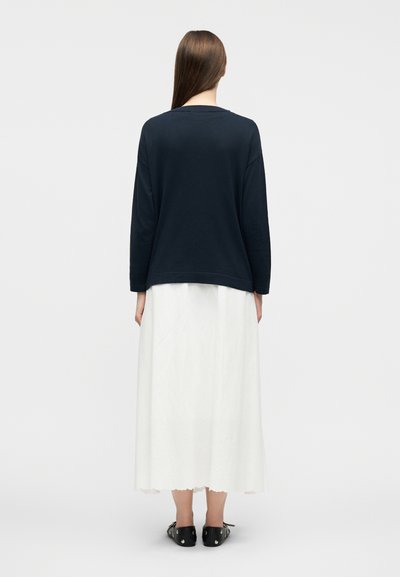 Γυναίκα με μακριά ίσια μαλλιά, φορώντας μαύρο μακρυμάνικο τοπ, άσπρη φούστα μέχρι τους αστράγαλους και μαύρα παπούτσια με τρουκς, γυρισμένη με την πλάτη.