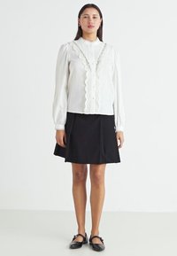 Blouse blanche à manches longues avec une bordure texturée et des boutons, associée à une jupe noire en ligne A et des chaussures plates noires. Design simple et élégant.