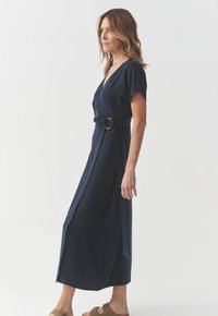 Abito avvolgente blu navy con scollatura a V, maniche corte e cintura con anello dorato. Tessuto morbido con lunghezza midi fluida e spacchi laterali.