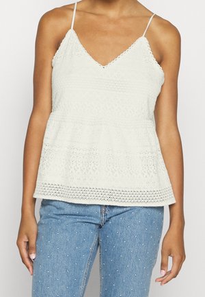 Top peplum blanco texturizado con escote en V y tirantes finos, que presenta intrincados patrones de crochet a lo largo del escote y el dobladillo. Combinado con vaqueros de mezclilla azules.