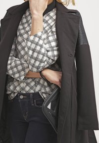 Kobieta w czarnym płaszczu, nałożonym na czarno-białą koszulę w kratę, trzymająca czarną torebkę, ubrana w ciemnoniebieskie dżinsy, na jednolitym tle.