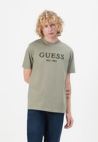 Maslinasto zelena pamučna majica s natpisom "GUESS EST. 1981" otisnutim tamnozelenim slovima. Kratki rukavi, okrugli izrez, opušteni kroj. Uparen s plavim trapericama.