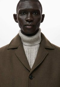 Abrigo de lana marrón con cuello con muesca sobre un suéter de cuello alto acanalado de color gris claro. Presenta un solo botón negro como detalle.