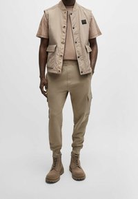 Beige ärmlös väst med framfickor, ljusbeige t-shirt, beige cargobyxor och bruna snörboots med texturerade sulor.