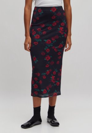 Frau trägt einen eng anliegenden schwarzen Midirock mit roten Rosenmustern, schwarze Socken, schwarze Ballerinas und ein weißes Oberteil vor einem einfarbigen Hintergrund.