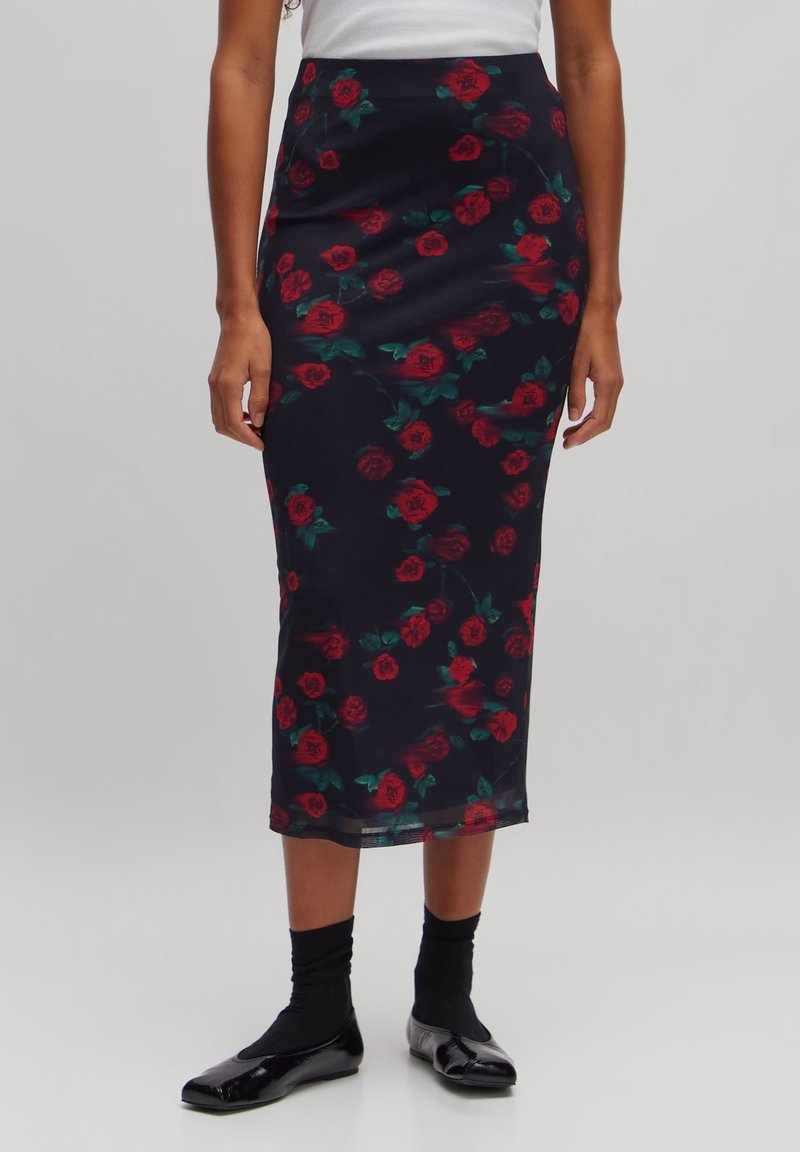 Femme portant une jupe midi noire ajustée avec des motifs de roses rouges, des chaussettes noires, des ballerines noires et un haut blanc sur un fond uni.