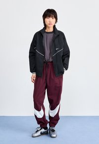 Svart Nike-jakke med hvite detaljer, gråstripete skjorte, maroon bukser med hvit Nike-logo og grå treningssko.