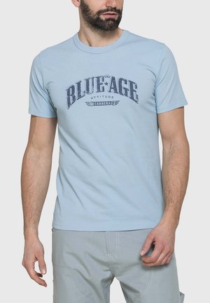 Uomo che indossa una maglietta azzurro chiaro con la scritta "THE BLUE AGE ATTITUDE CARRERA" e pantaloni grigi, in piedi davanti a uno sfondo semplice.