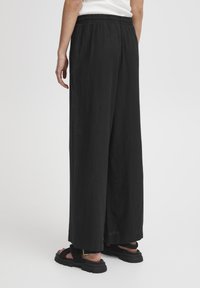 Melnas platās bikses, izgatavotas no viegla auduma, ar elastīgu jostu un gludu tekstūru. Valkātas ar melniem siksnu sandaliem.