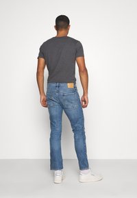 Calças de jeans azul para homem com um corte reto, apresentando um acabamento texturizado, dois bolsos traseiros e um rótulo da marca castanho na cintura.