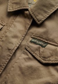Bruine herringbone overhemd met een kraag, voorzien van patchzakken en een groene "SUPERDRY" label geborduurd op de linker zak.