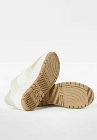 Bílé tenisky s hladkým svrškem a béžovou gumovou podrážkou. Mají kulatou špičku, texturovanou podrážku pro dobrou trakci a minimalistický design.