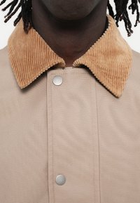 Veste beige avec un col en velours côtelé. Le col est beige avec une texture rainurée, fixé par un bouton-pression argenté à l'avant.