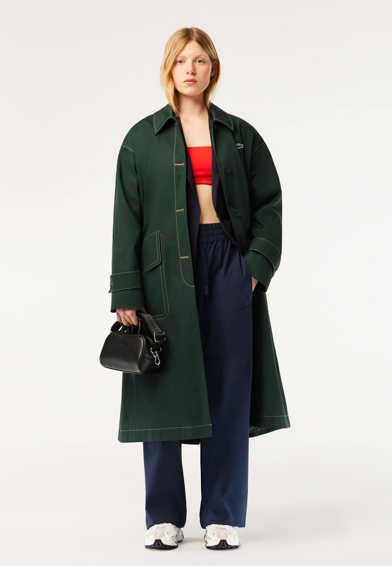 Long manteau vert avec des coutures jaunes, crop top rouge, pantalon bleu marine et sac à main noir. Le modèle porte des baskets blanches avec des accents violets.