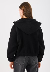 Sort fleecejakke med hette, som har en strukturert overflate, avslappet passform og senkede skuldre. Brukt med grå denimbukser.
