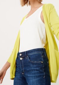 Světle žlutý cardigan přes bílý top bez rukávů, v kombinaci s tmavě modrými džíny se zdvojeným zapínáním a viditelnými švy.