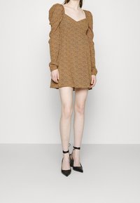 Mini-robe marron à motifs, manches longues bouffantes, encolure carrée et taille froncée, associée à des talons noirs à bout pointu.