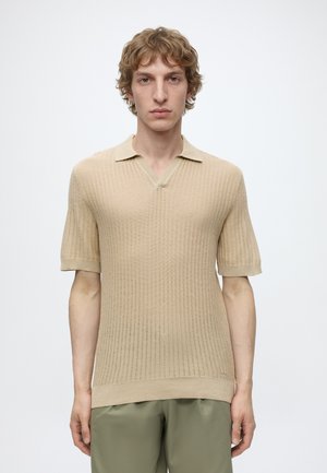 Jeune homme aux cheveux bouclés portant un polo en maille côtelée beige à manches courtes et un pantalon vert olive, debout sur un fond blanc uni.